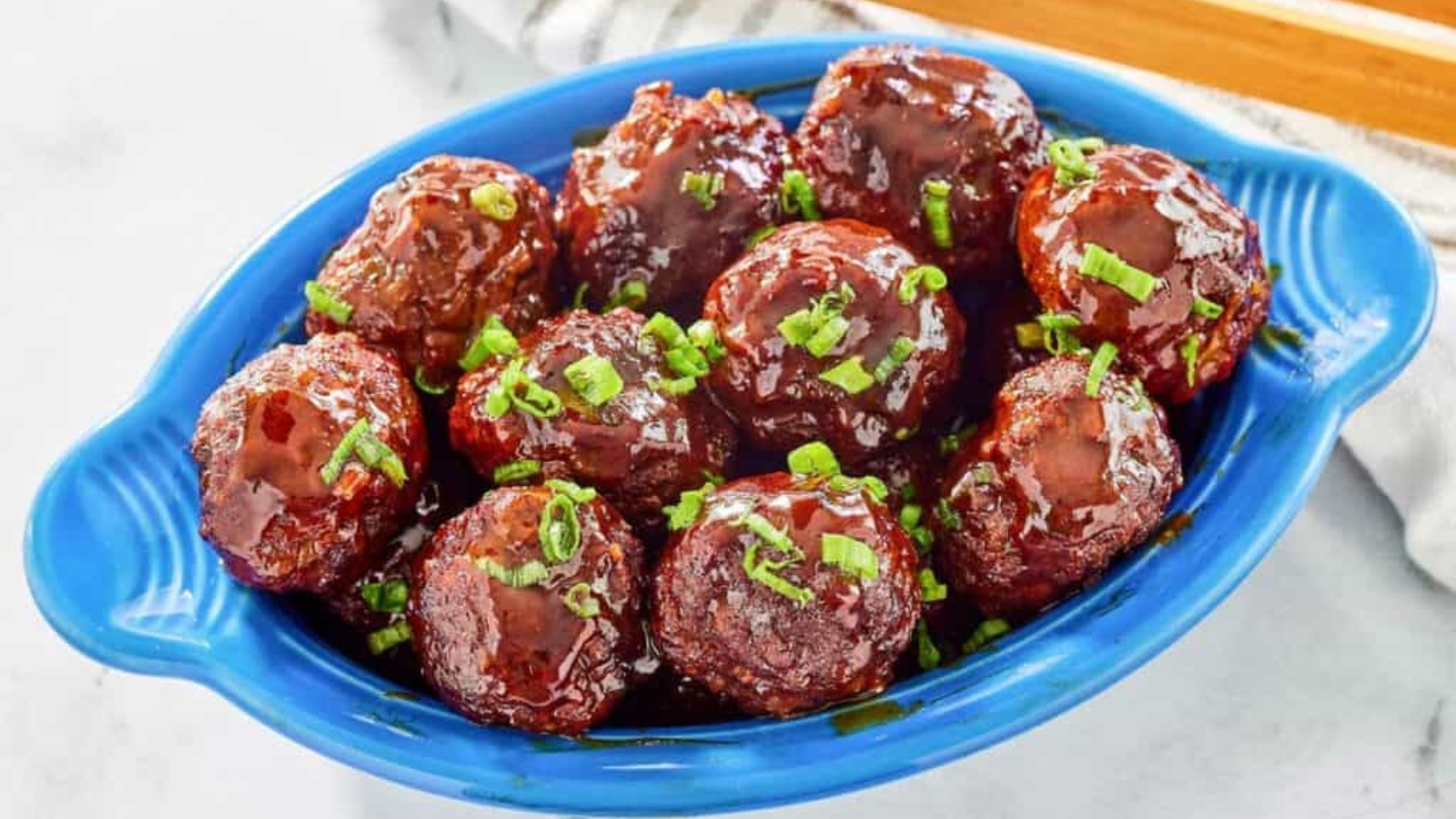 Meatballs coated in a glossy sauce, garnished with chopped green onions, served in a blue dish.