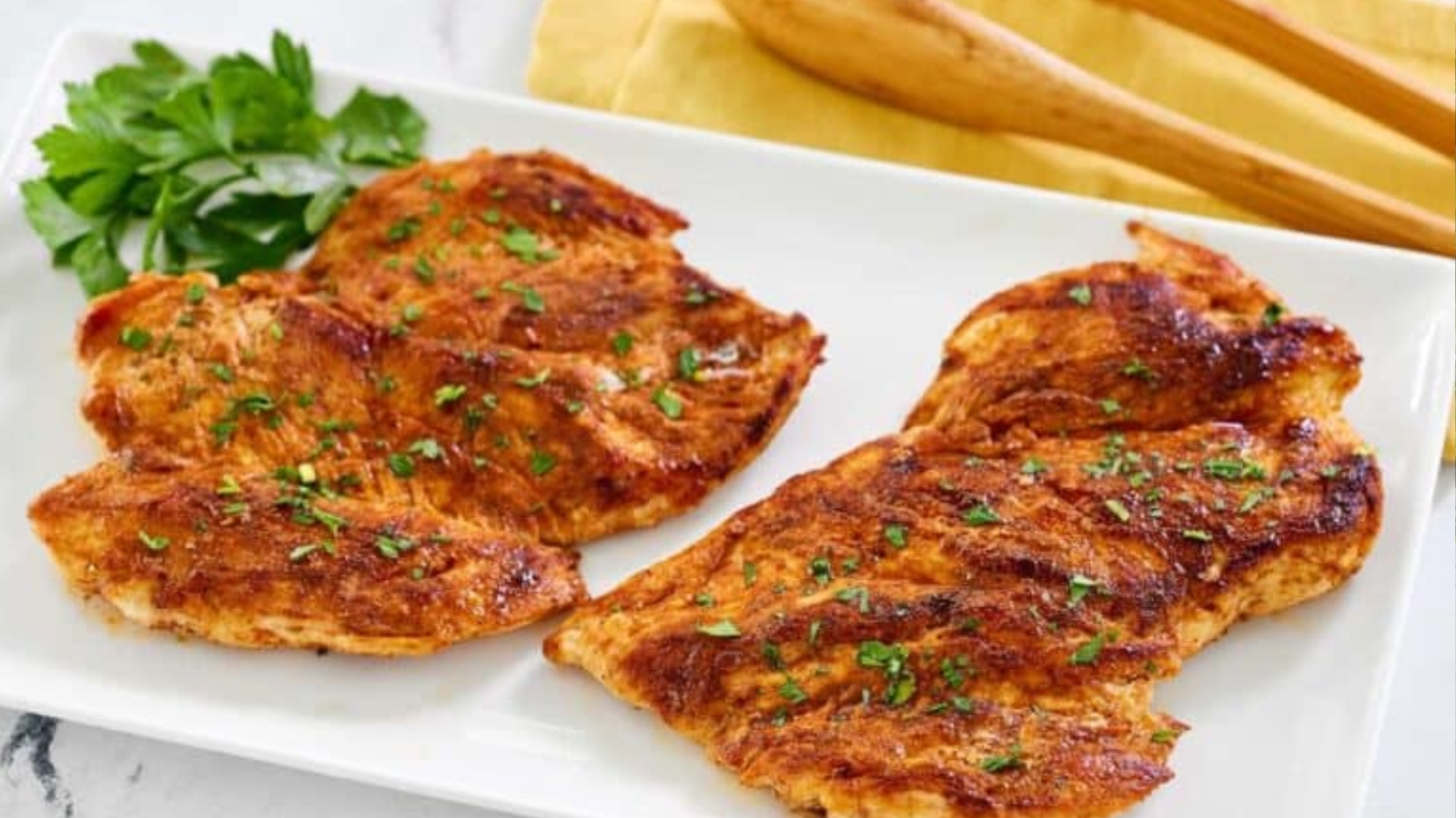 Grilled chicken breasts with seasoning, garnished with herbs on a white plate with parsley and wooden utensils.