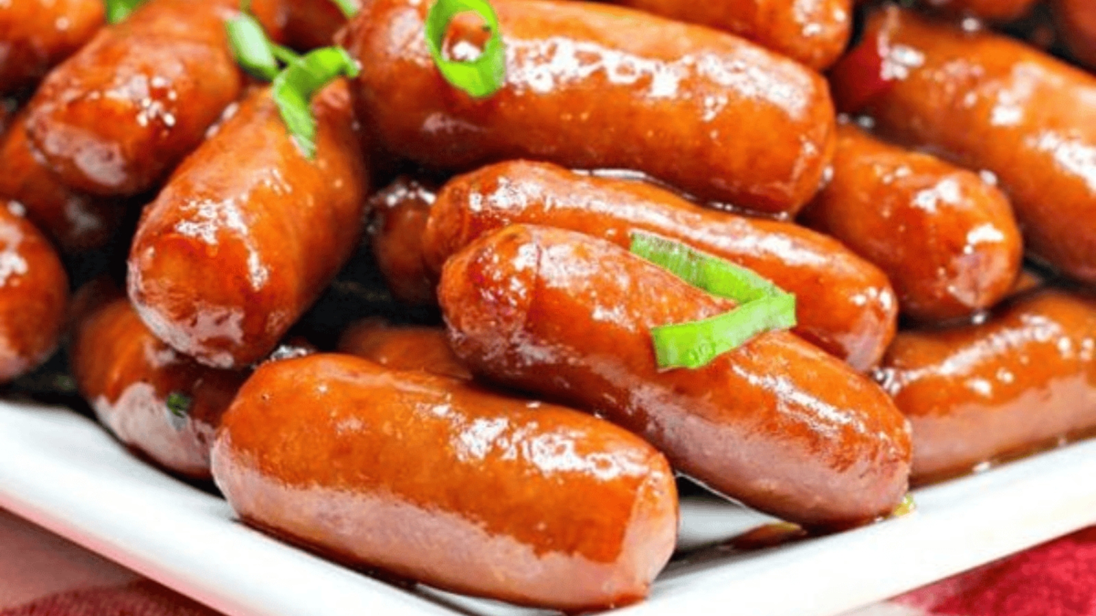 Cocktail sausages glazed with sauce, garnished with sliced green onions, served on a white platter.