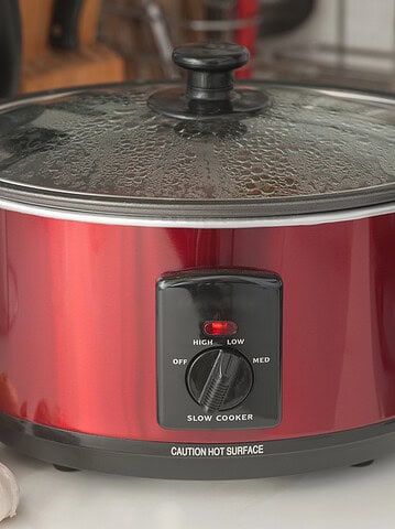 A red slow cooker with a glass lid sits on a kitchen counter near garlic, onions, and peppers, ready to help you avoid common slow cooker mistakes in your next flavorful meal.
