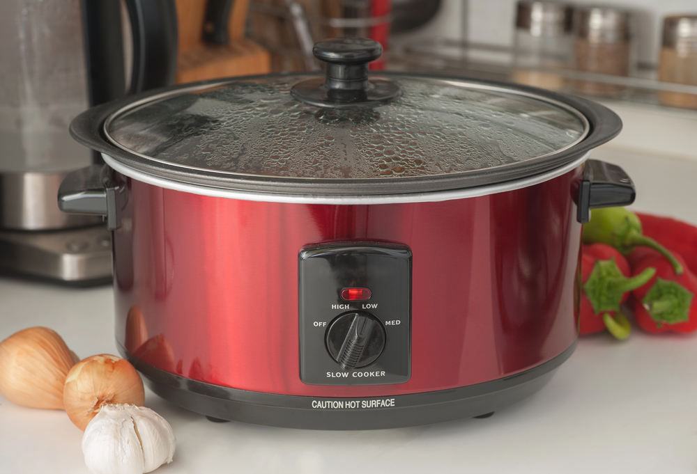 Red slow cooker on a kitchen counter with onions, garlic, and a bell pepper nearby&mdash;a perfect setup when you want to avoid common slow cooker mistakes and enjoy easy, delicious meals.