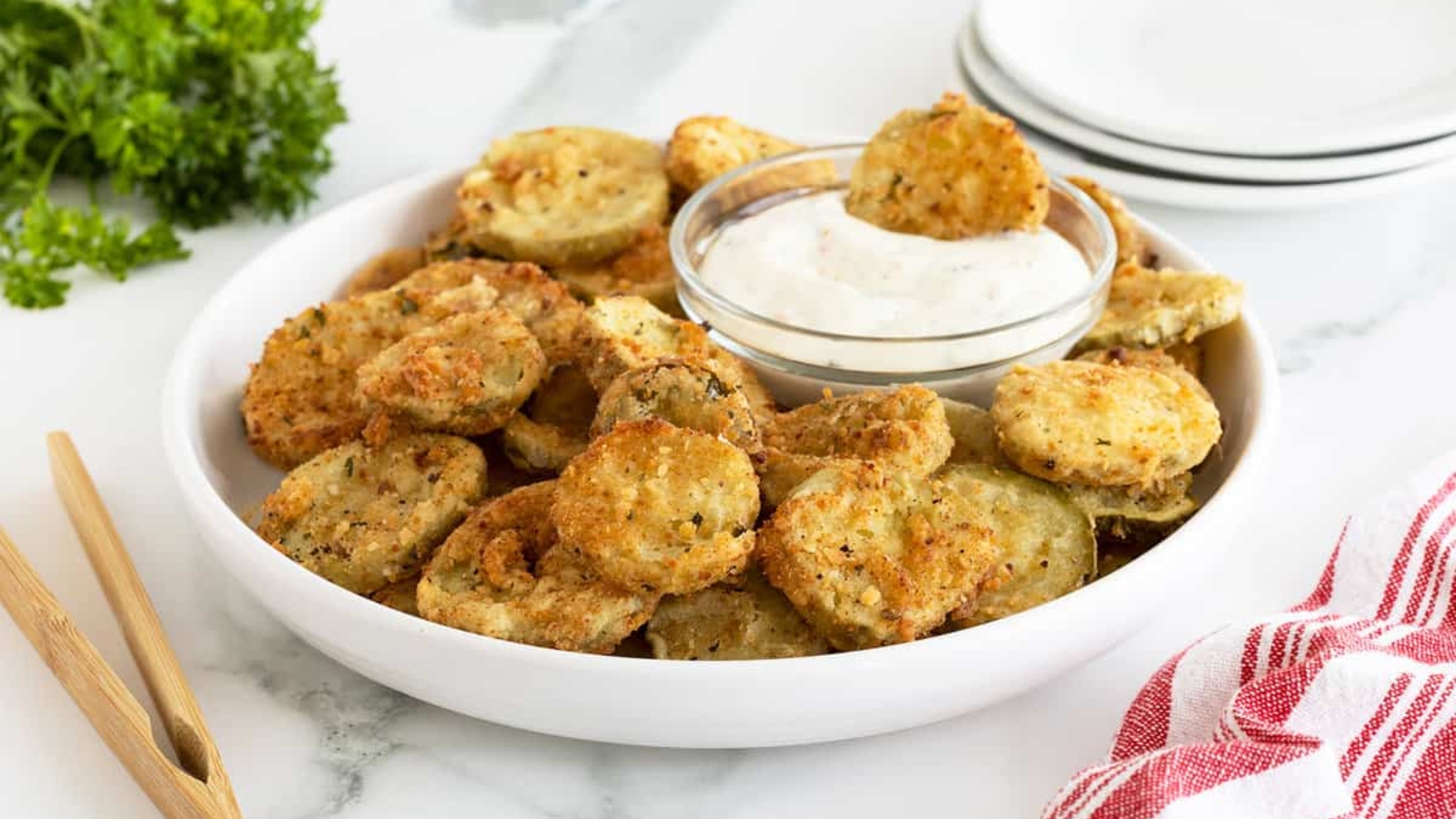 A plate of fried pickles with a bowl of dipping sauce sits on a table next to a red-striped towel.