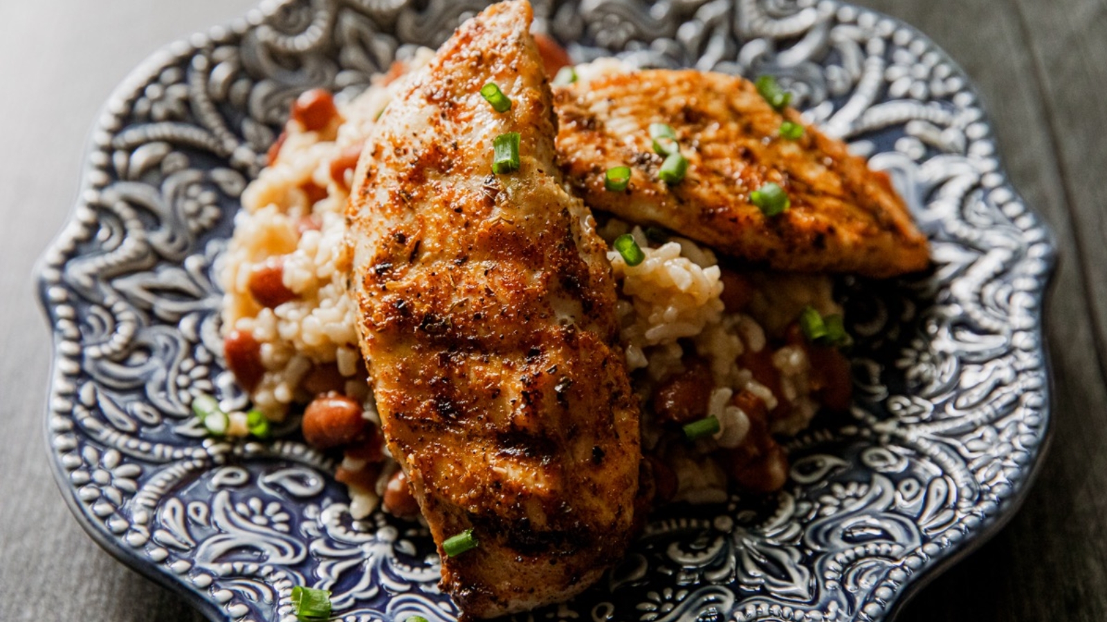 Grilled chicken breast served on rice and beans, garnished with chopped green onions on a blue patterned plate.