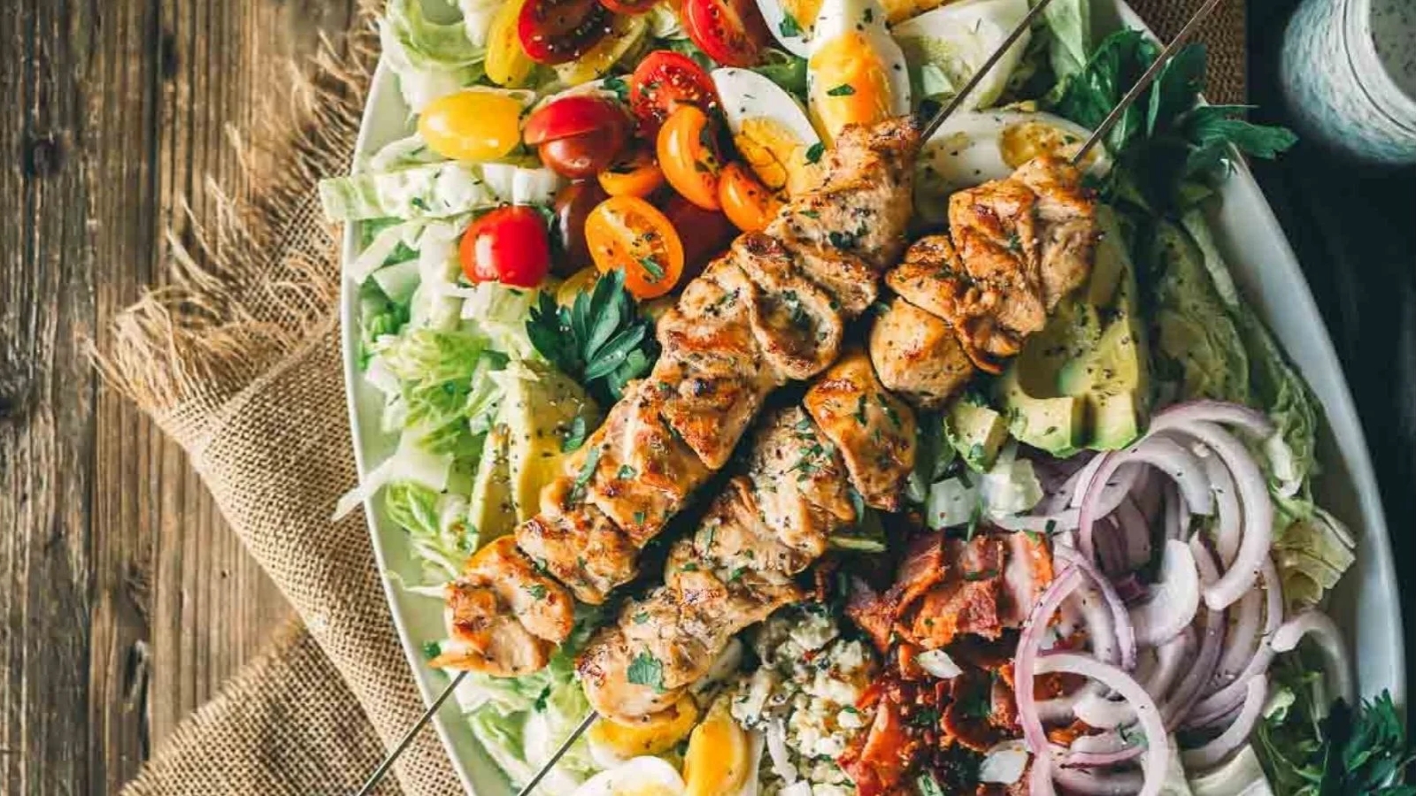Grilled chicken skewers on a salad with tomatoes, avocado, red onions, lettuce, bacon, and cheese.