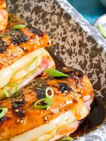 Three baked chicken breasts stuffed with cheese and ham, topped with sliced green onions in a speckled baking dish.