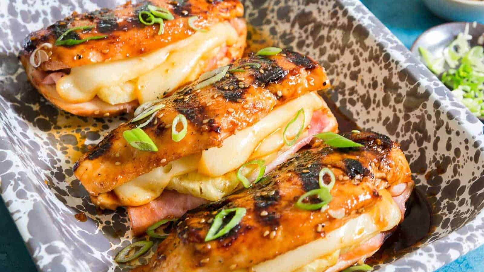 Three grilled chicken breasts stuffed with ham and melted cheese are artfully arranged in a speckled ceramic dish, making them the perfect backyard BBQ hero. Garnished with sliced green onions and glazed with a savory sauce, this grilled meal invites everyone to the table.