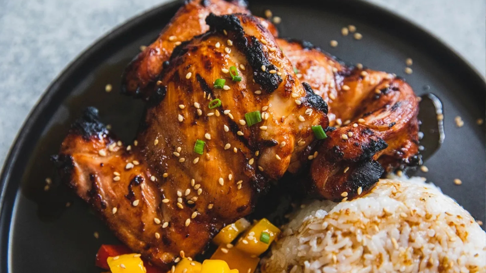 Grilled chicken with sesame seeds, served with white rice and colorful diced vegetables on a black plate.