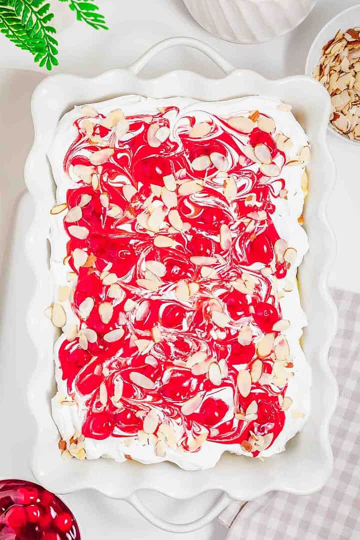 Rectangular heaven on earth cake with whipped topping, red swirls, and sliced almonds in a white baking dish.