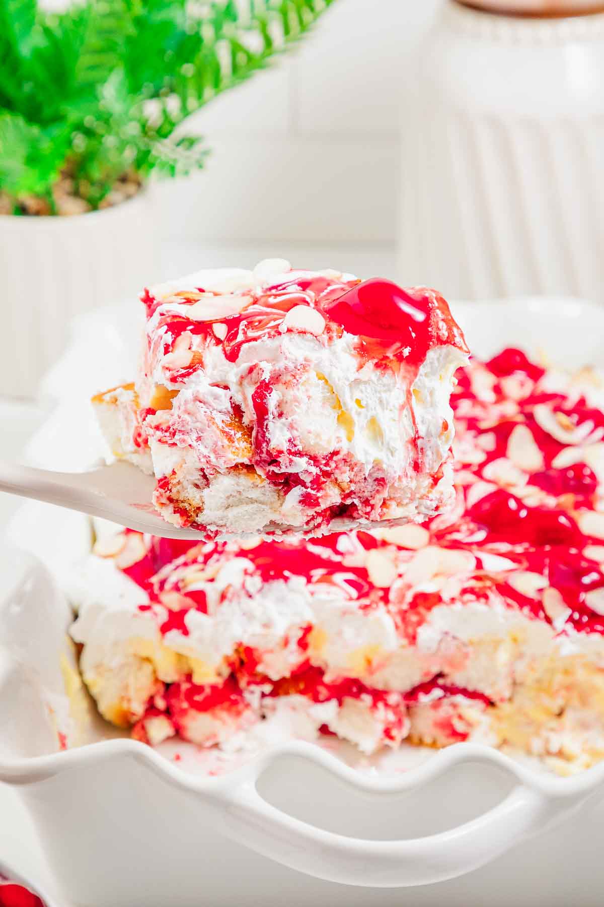 A spatula lifts a slice of heaven on earth cake, a cherry dessert topped with whipped cream and almond slices, from a baking dish.