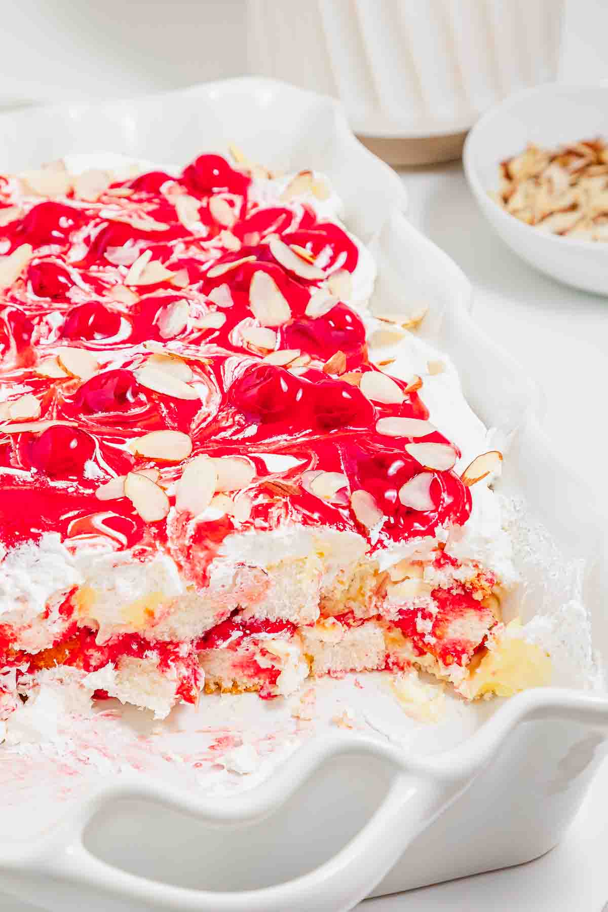 Cherry-topped heaven on earth cake layered with whipped cream and almond slices in a white dish, partially served.