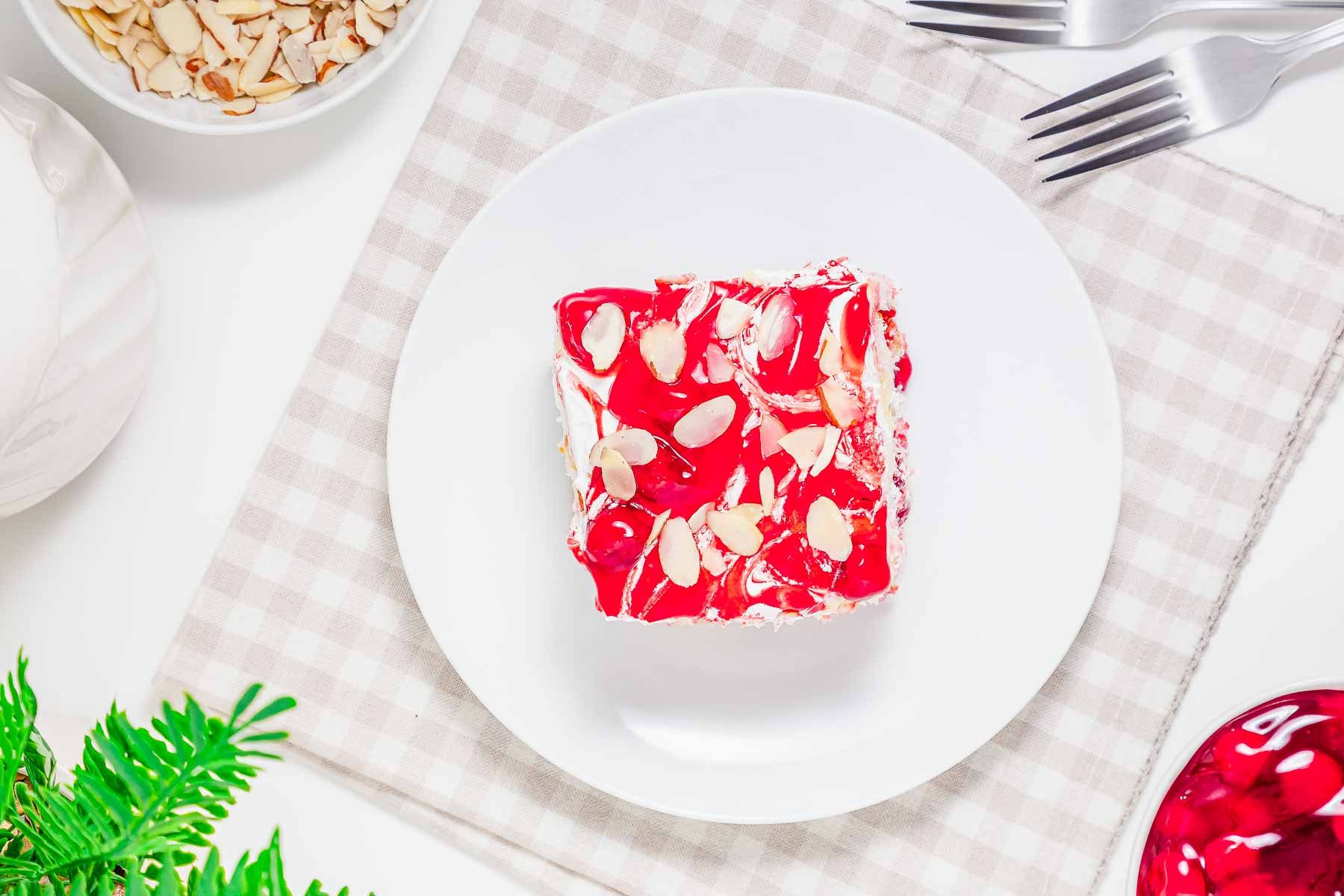 A square slice of cherry dessert, reminiscent of heaven on earth cake, is topped with almond slices on a white plate, set on a checkered cloth.