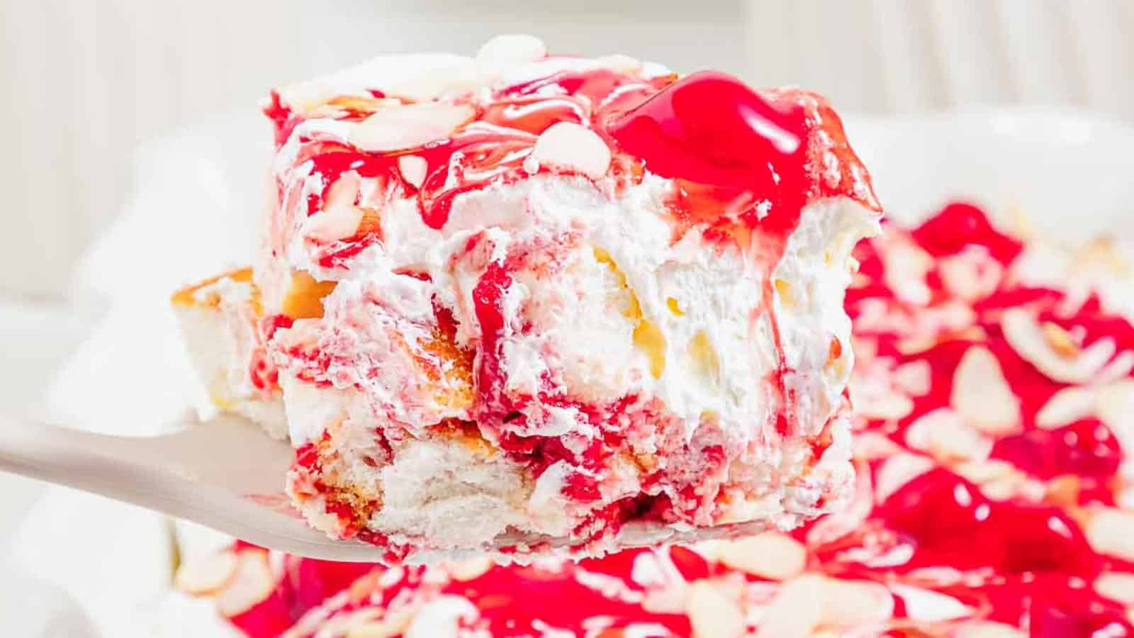 A close-up of a cherry almond dessert with whipped cream, cherry topping, and sliced almonds on a spatula.