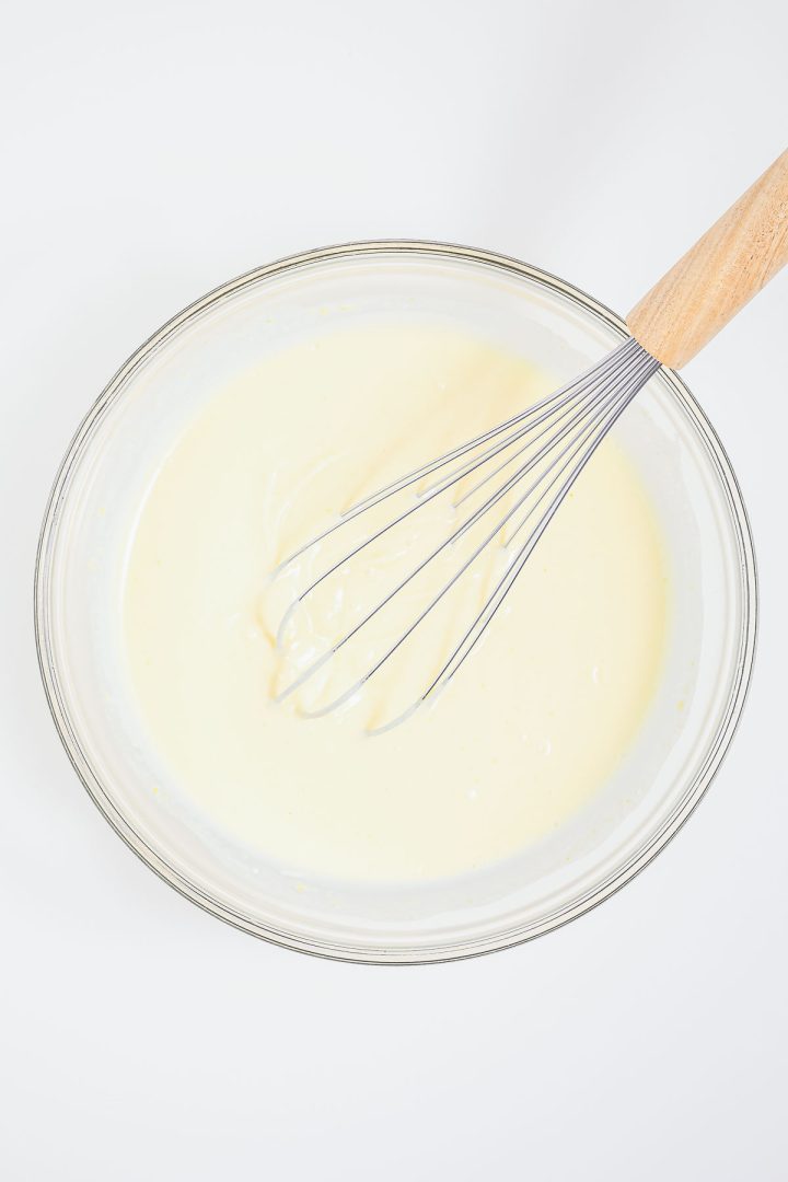A whisk in a clear bowl of pale, creamy batter—perfect for making a heaven on earth cake—on a white background.