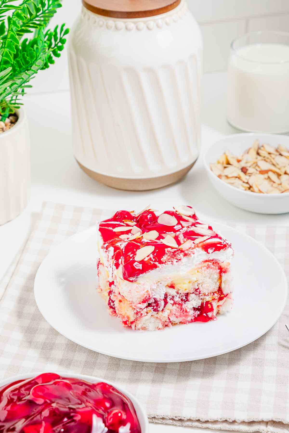 A slice of cherry dessert with cream and almonds on a white plate, reminiscent of heaven on earth cake, is surrounded by bowls and a glass of milk.