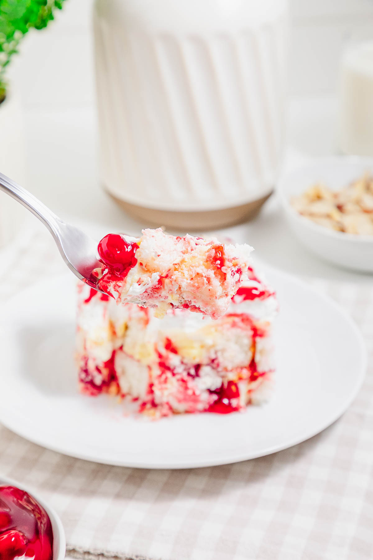 A fork holds a bite of cherry cake with whipped cream, above a plate with a larger piece of the heaven on earth cake.