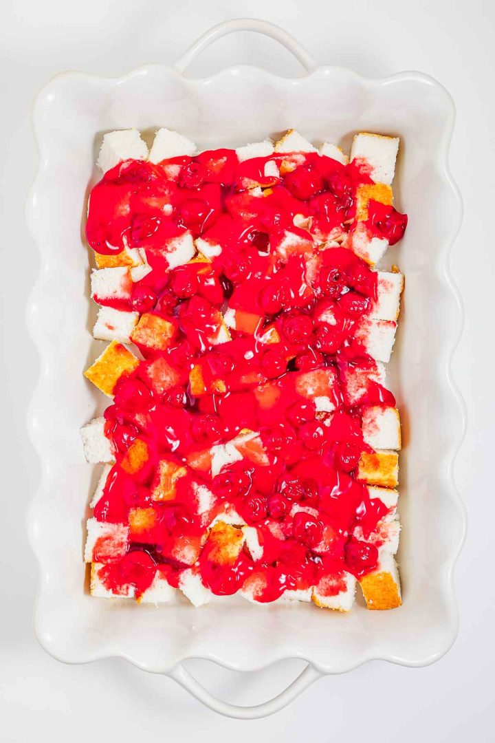 Cubed heaven on earth cake topped with bright red cherry pie filling in a white scalloped baking dish.