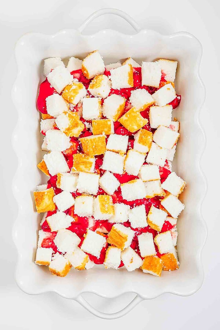 Cubed cake pieces layered over bright red fruit filling in a white scalloped baking dish create a classic heaven on earth cake.