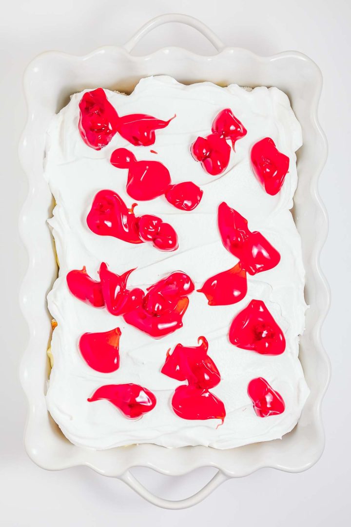 A white frosted heaven on earth cake sits in a baking dish, with dollops of bright red topping scattered on top.