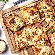 Rectangular pieces of cheesy Italian Sausage Sheet Pan Pizza with pepperoni and sausage are arranged on a wooden board, a pizza cutter nearby.