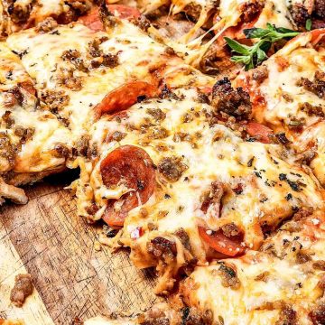 Sliced pizza on a wooden board, topped with pepperoni, Italian sausage, melted cheese, and herbs&mdash;just like a classic Italian Sausage Sheet Pan Pizza.
