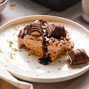 Square Kinder Bueno No Bake Cake with chocolate drizzle, creamy filling, and cookie base on a white plate with crumbs.