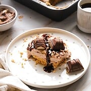 A Kinder Bueno No Bake Cake topped with chocolate syrup and candy sits on a plate, with a baking pan and coffee nearby.
