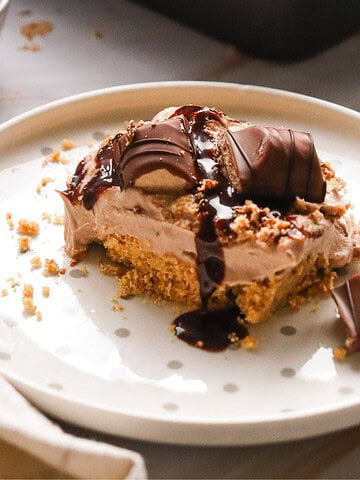 Square Kinder Bueno No Bake Cake with chocolate drizzle, creamy filling, and cookie base on a white plate with crumbs.