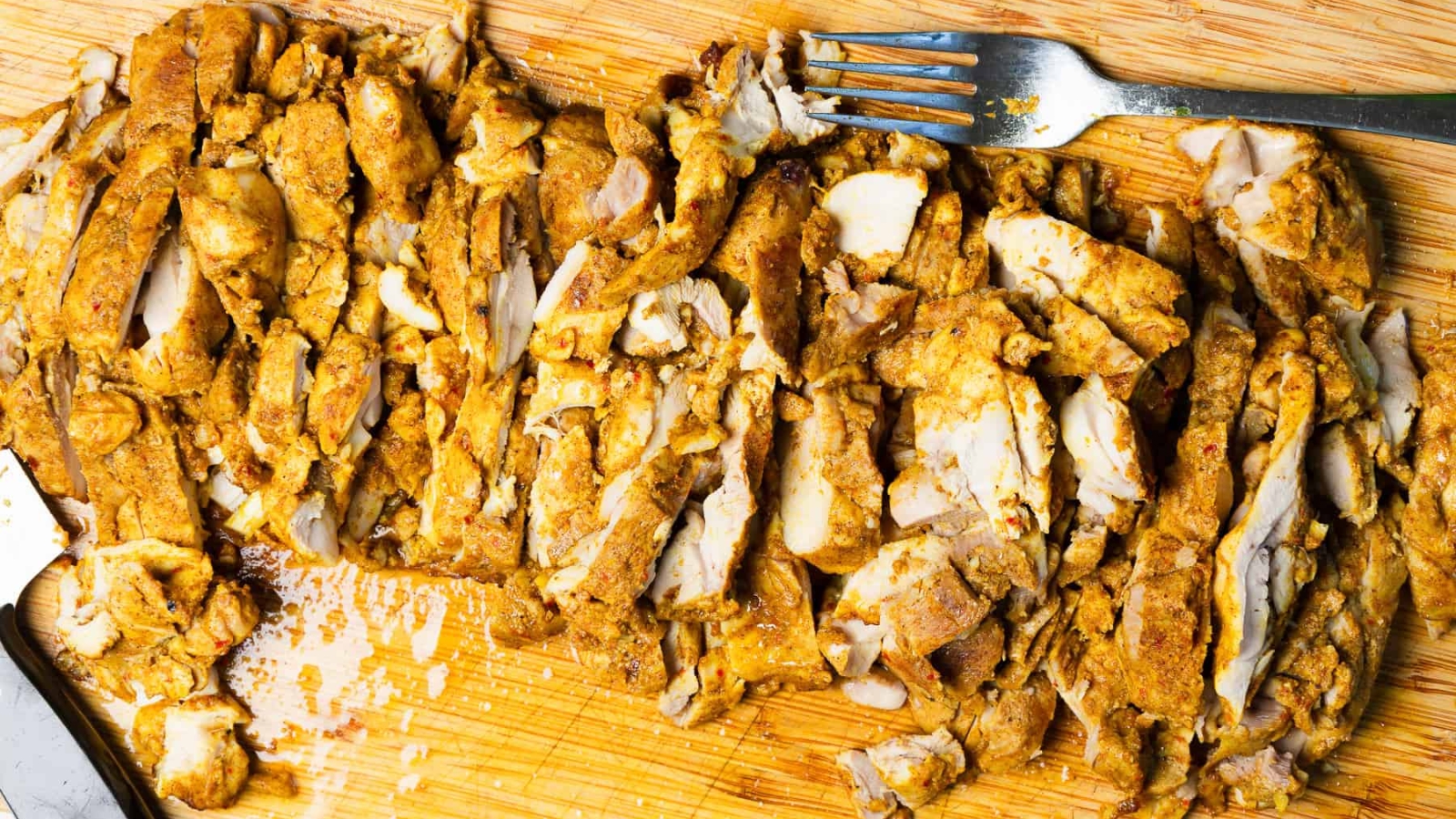 Sliced, cooked seasoned chicken on a wooden cutting board with a fork in the background.