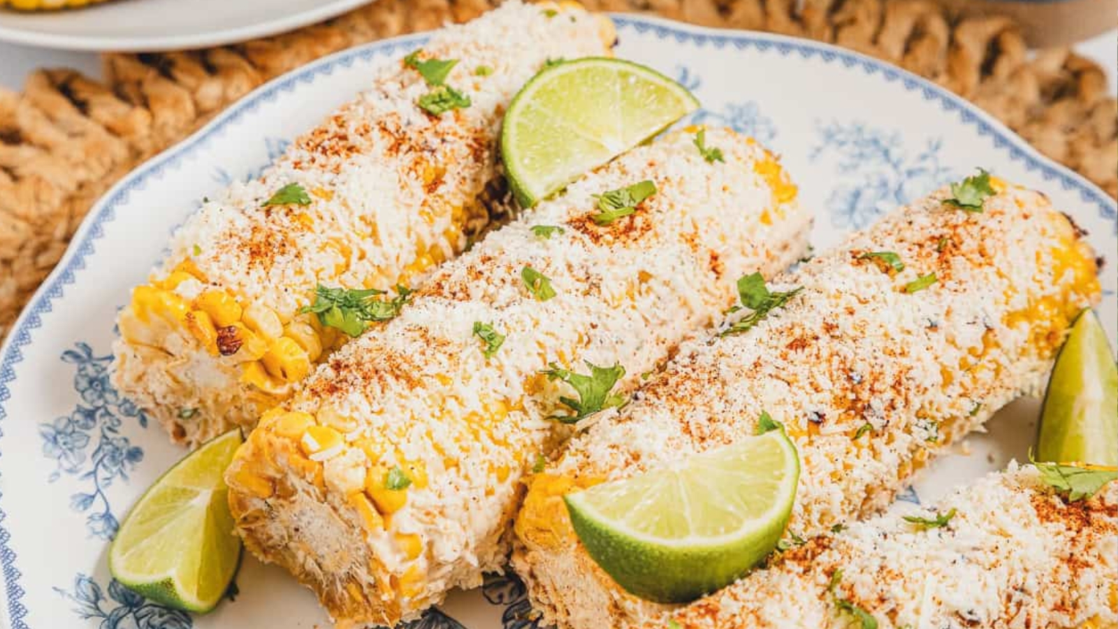 Three elotes (Mexican street corn) on a plate, topped with cheese, chili powder, and lime wedges.