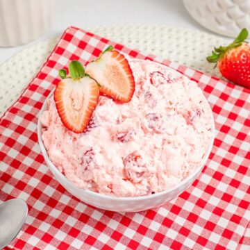 A bowl of Strawberry Fluff salad with sliced strawberries on top, placed on a red checkered cloth.