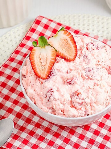 A bowl of Strawberry Fluff salad with sliced strawberries on top, placed on a red checkered cloth.