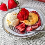 A plate with strawberry cobbler, vanilla ice cream, and fresh strawberries, reminiscent of a Strawberry Shortcake Casserole, with more cobbler in the background.