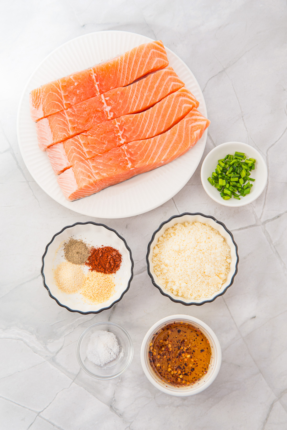 Four raw salmon fillets, green onions, breadcrumbs, spices, salt, and a bowl of oil are arranged on a marble surface—perfect ingredients for making flavorful Air Fryer Parmesan Salmon Bites.