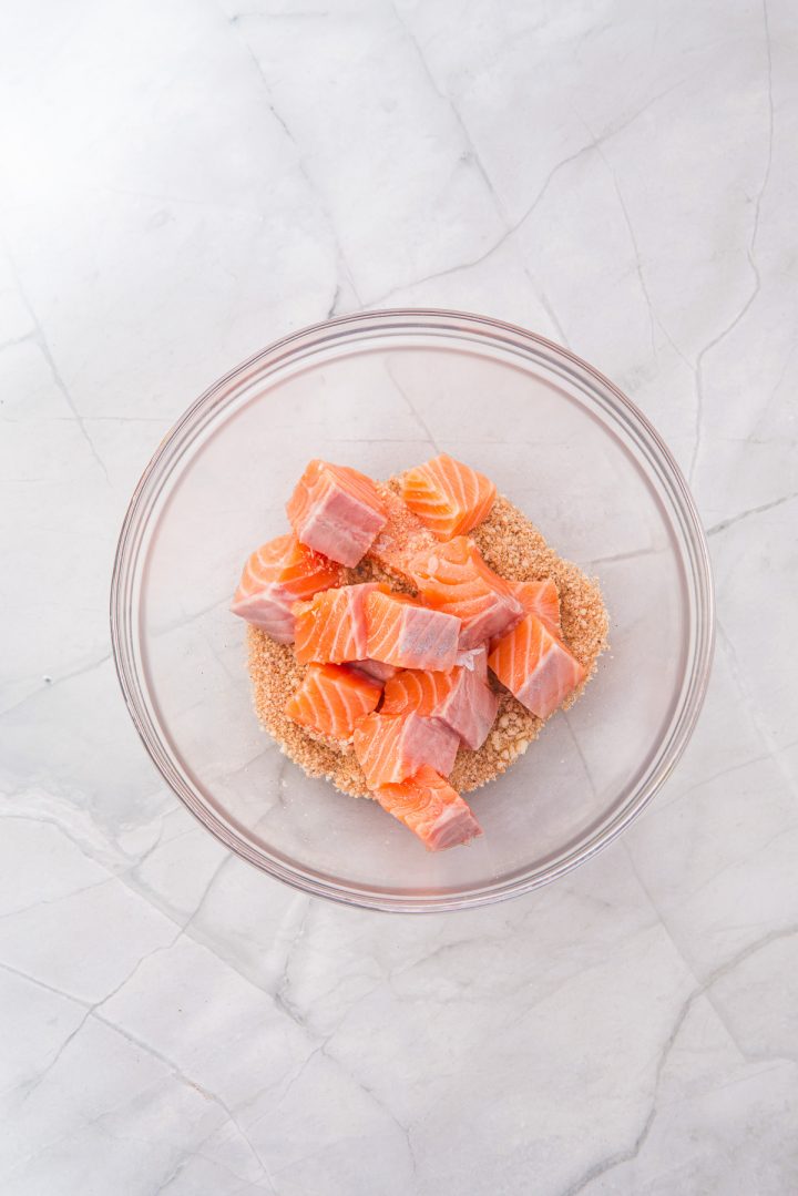 Cubed raw salmon, perfect for making Air Fryer Parmesan Salmon Bites, sits on a layer of brown sugar in a clear glass bowl on a marble countertop.