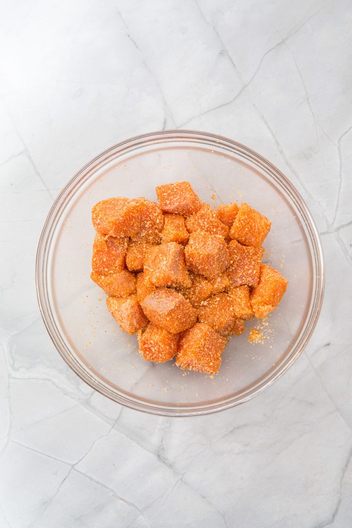 Cubed sweet potatoes coated with seasoning in a clear glass bowl on a light marble surface, perfect as a flavorful side dish to serve with Air Fryer Parmesan Salmon Bites.
