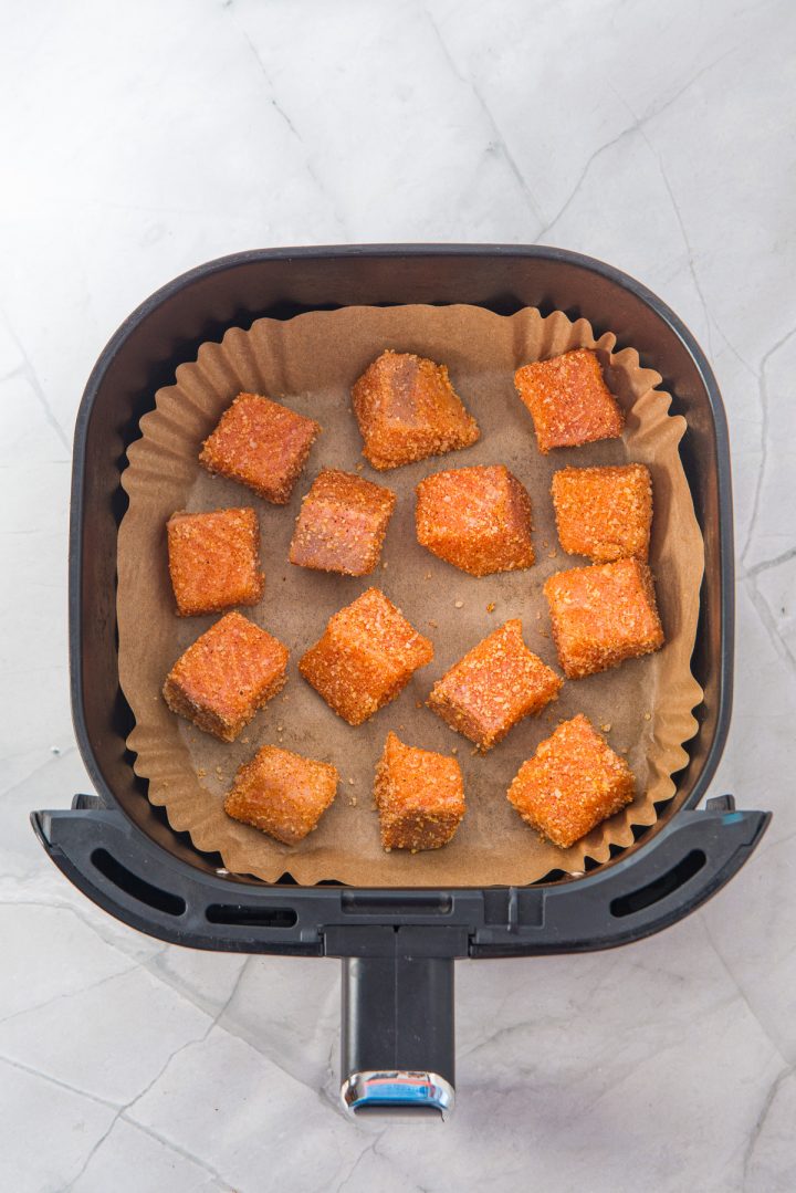 Air fryer basket with parchment paper holding twelve crispy Air Fryer Parmesan Salmon Bites on a marble surface.