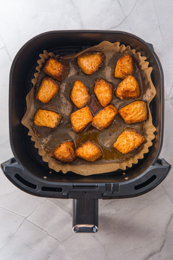Crispy golden potato wedges in parchment paper sit beside delicious Air Fryer Parmesan Salmon Bites in an air fryer basket on a marble countertop.