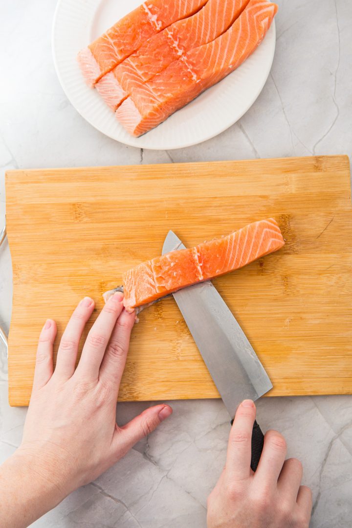 Hands slicing a raw salmon fillet on a wooden cutting board, preparing perfect portions for Air Fryer Parmesan Salmon Bites, with more salmon on a plate above.