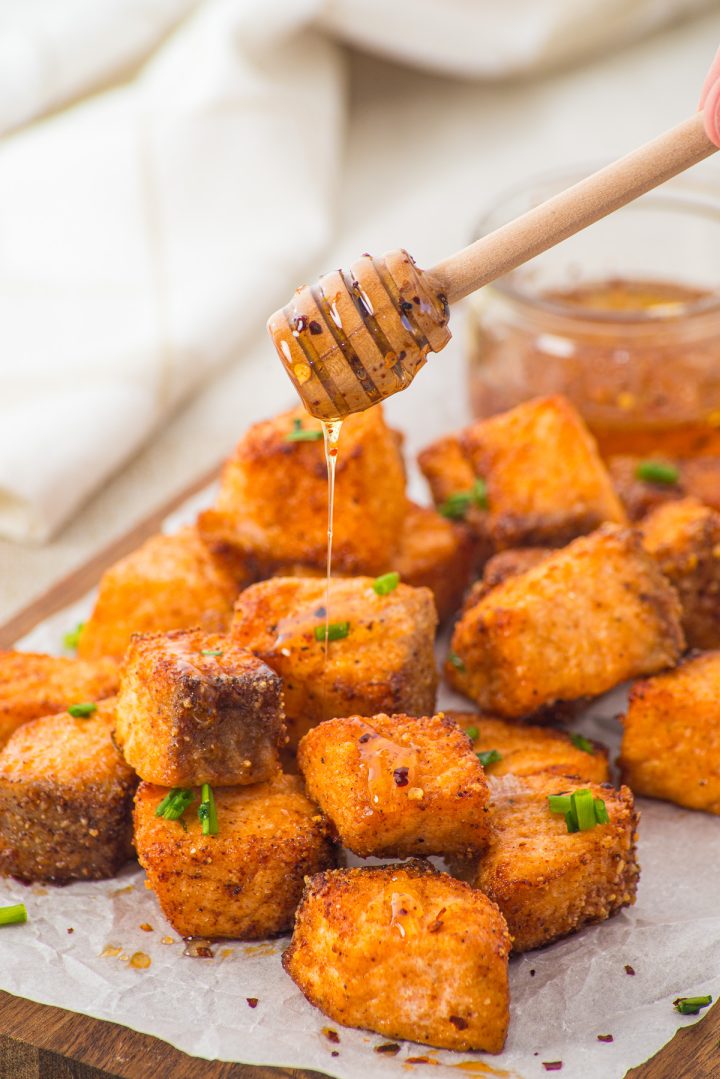Honey being drizzled over crispy, golden Air Fryer Parmesan Salmon Bites garnished with chives on parchment paper.