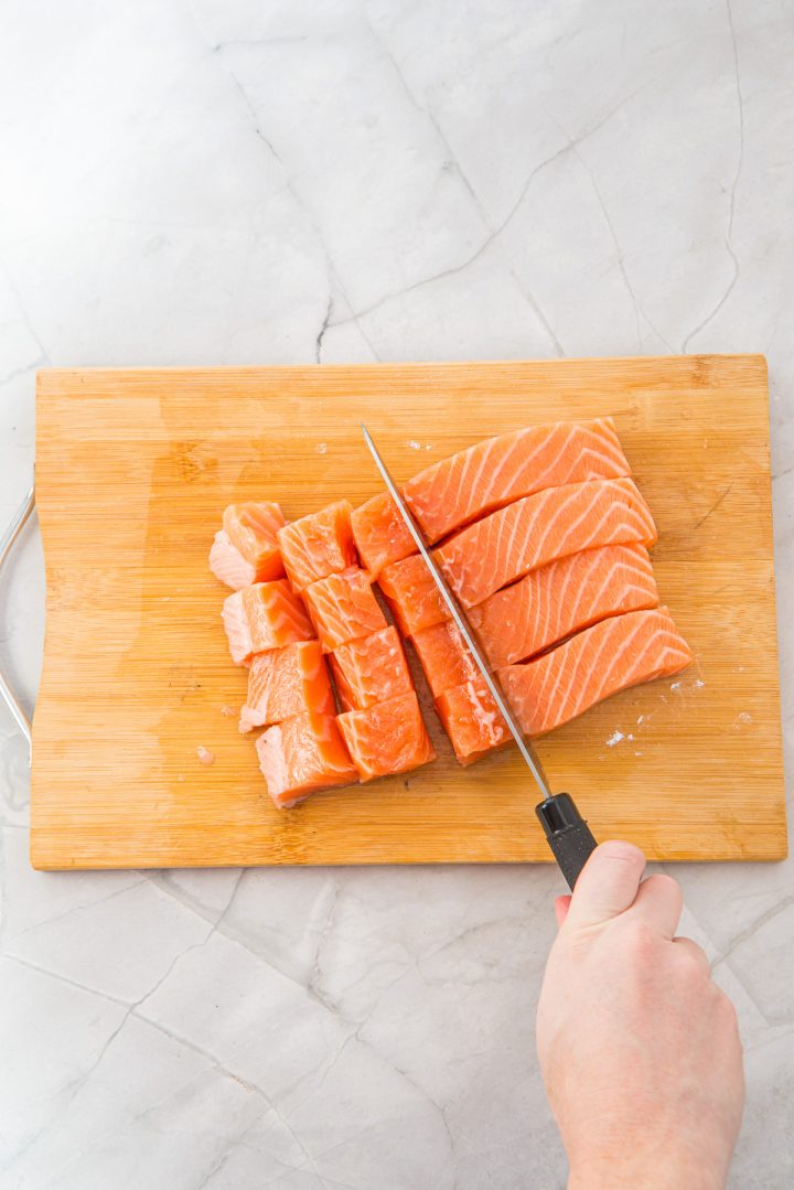 Hand slicing raw salmon into thick pieces on a wooden cutting board placed on a light-colored countertop, preparing the perfect base for delicious Air Fryer Parmesan Salmon Bites.