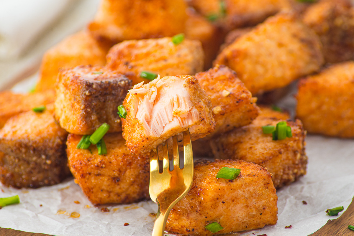 A golden fork holds a bite of seasoned, cooked Air Fryer Parmesan Salmon Bites garnished with chopped green onions.
