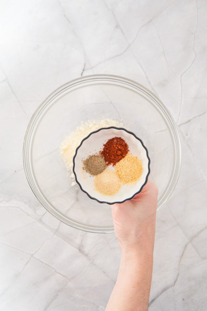 A hand holds a bowl of four spices over an empty glass mixing bowl on a marble countertop, preparing flavorful seasonings for Air Fryer Parmesan Salmon Bites.
