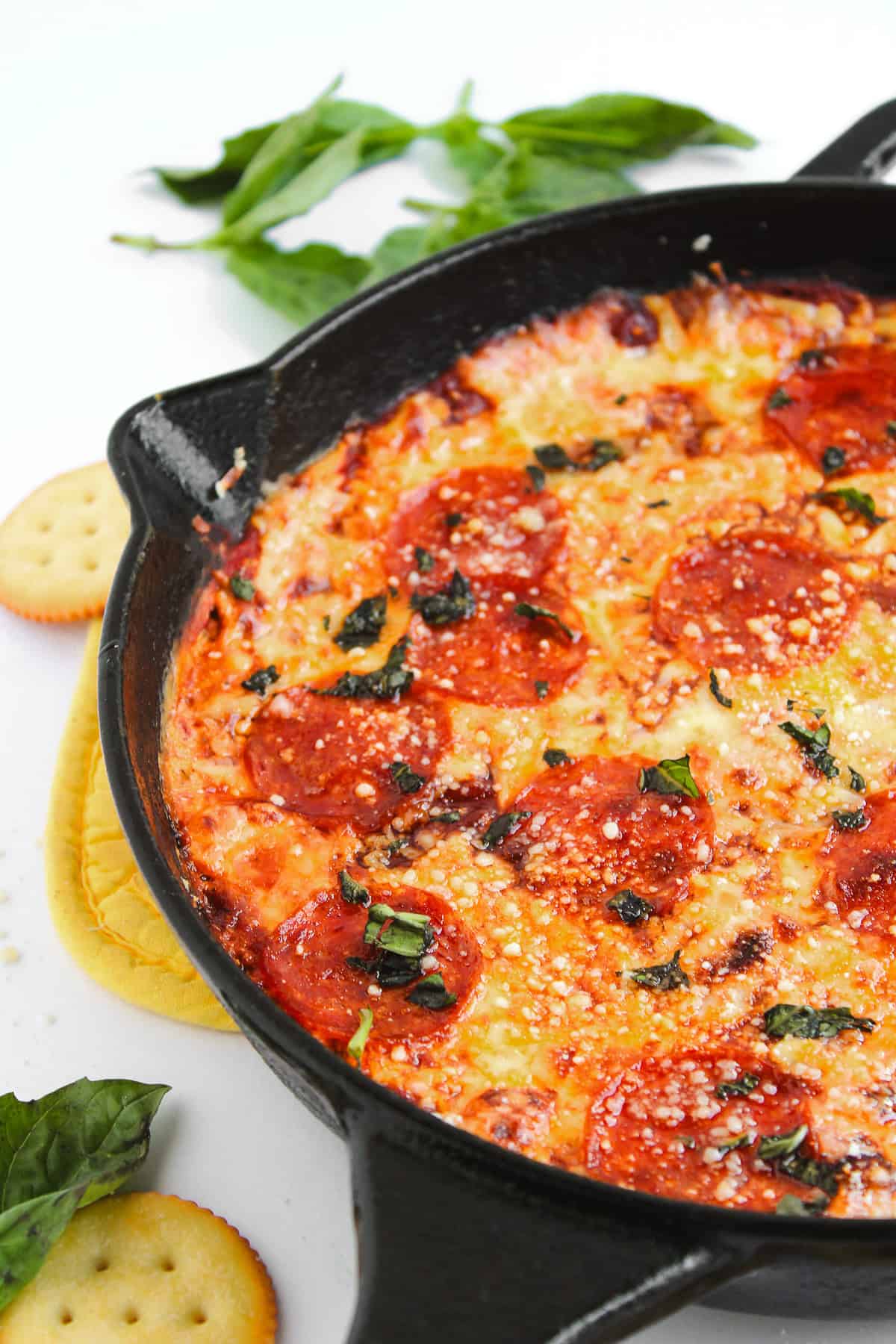 Skillet pepperoni pizza dip topped with cheese and basil, served hot with a drizzle of hot honey pizza dip, alongside crackers and fresh basil leaves.