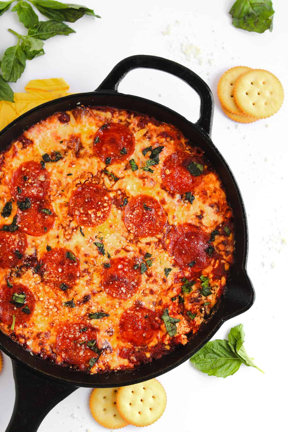 Cast iron skillet with hot honey pizza dip, pepperoni, basil, and crackers on a white surface with scattered herbs.