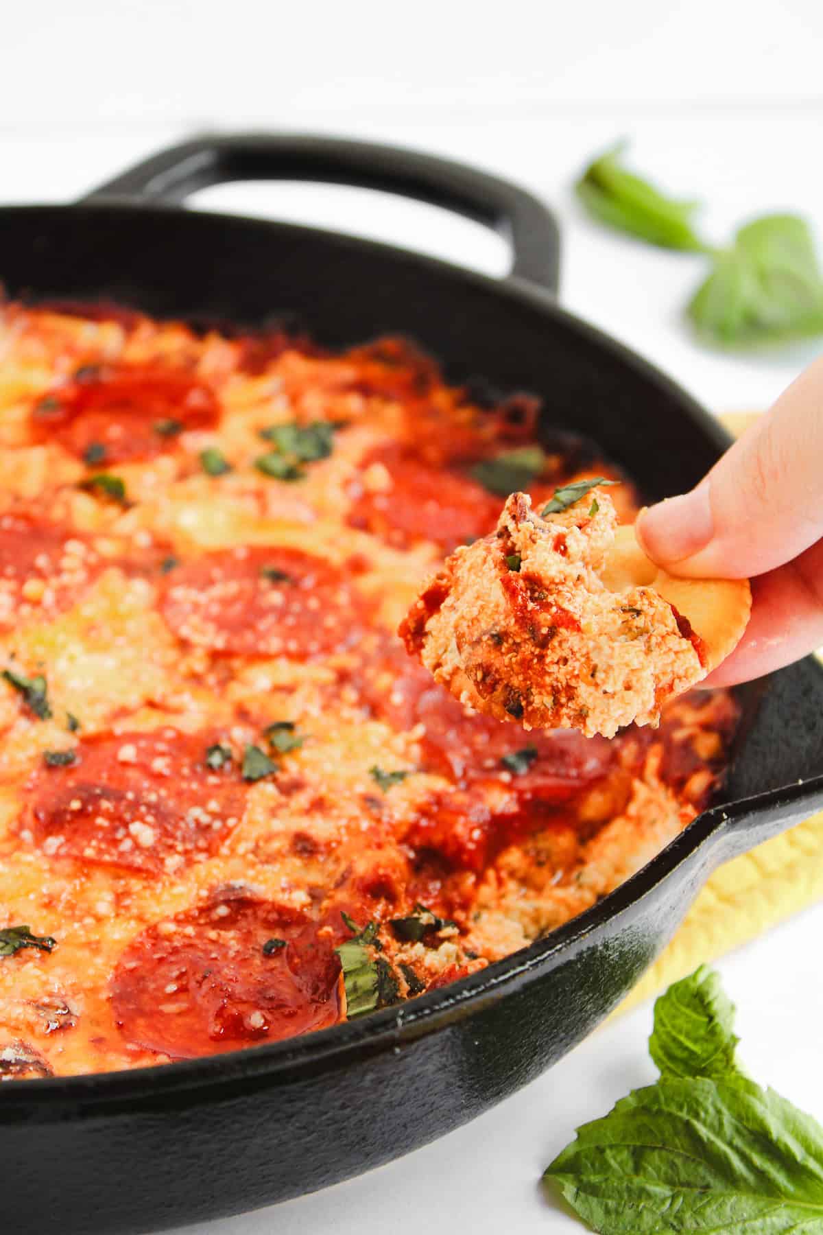 A hand holds a chip with cheesy, pepperoni hot honey pizza dip scooped from a cast iron skillet, topped with herbs.