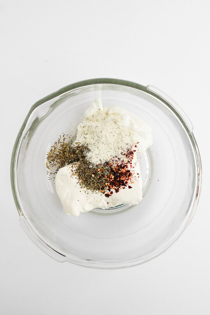 Glass bowl with sour cream, dried herbs, pepper, red pepper flakes, and a touch of hot honey pizza dip on a white background.