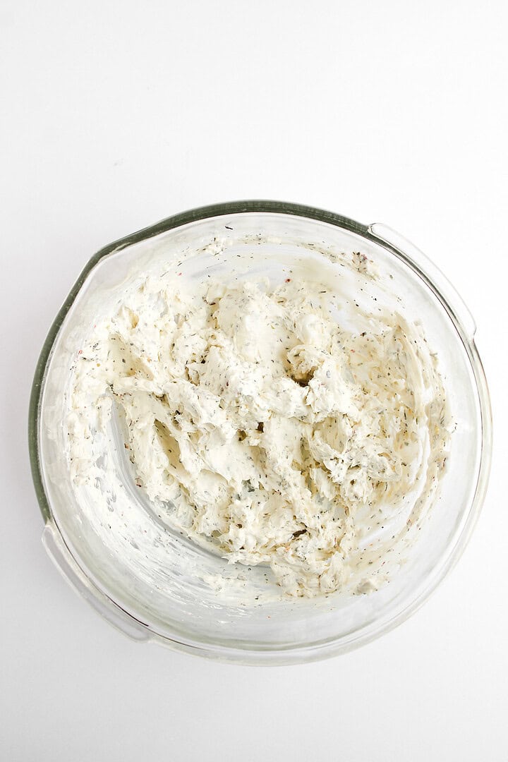Creamy, herbed cheese spread mixed in a clear glass bowl on a white background, perfect as a hot honey pizza dip.