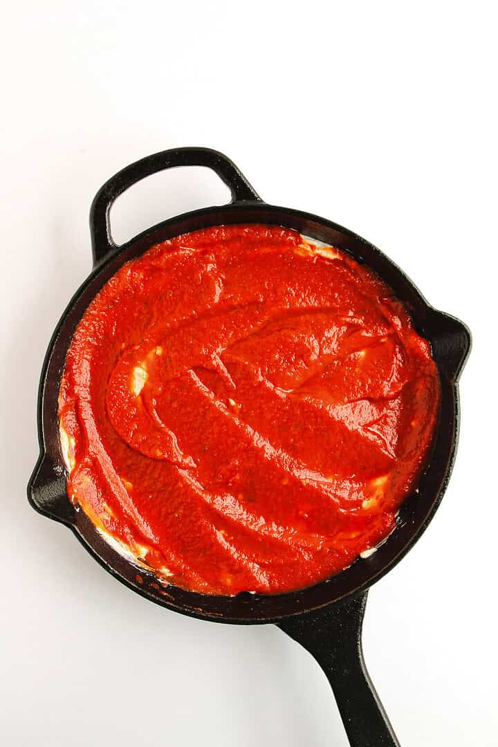 A black skillet filled with pizza topped with tomato sauce and served alongside a hot honey pizza dip, viewed from above on a white background.