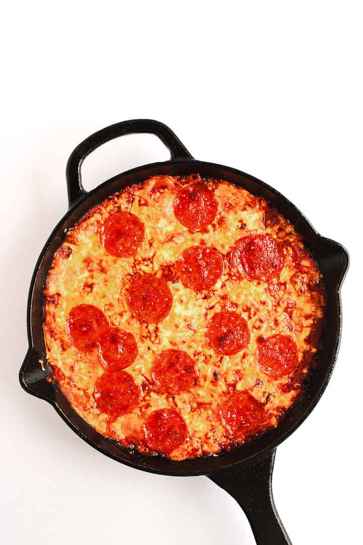 A pepperoni pizza baked in a black cast-iron skillet on a white background, served with a side of hot honey pizza dip.