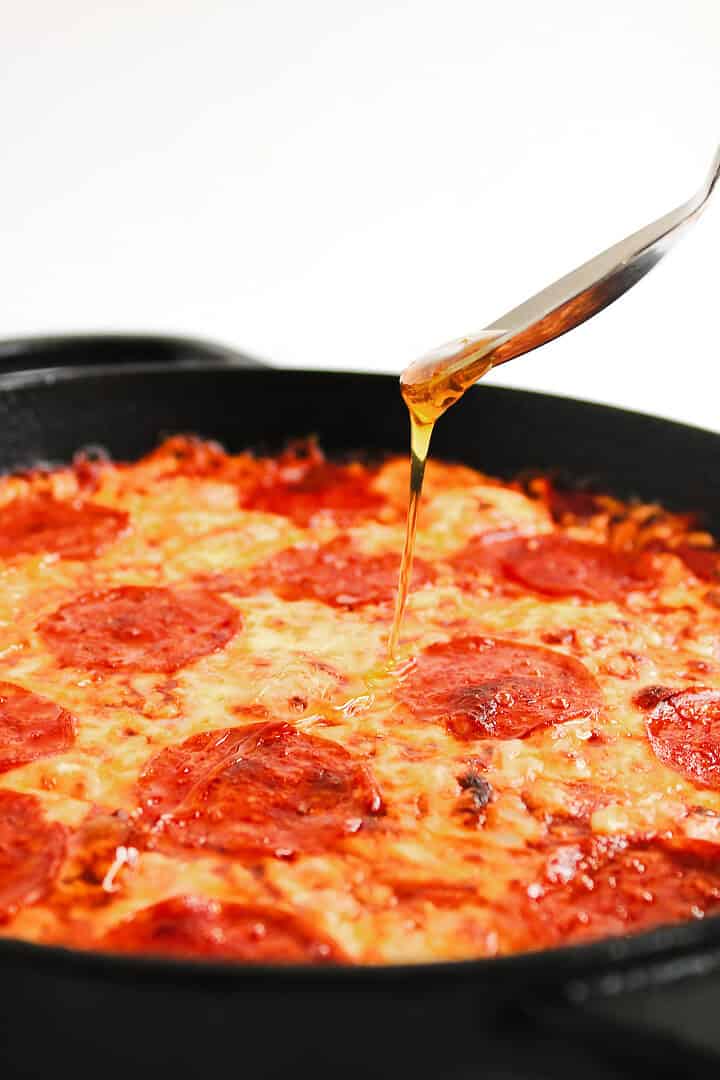 A spoon drizzling oil over a pepperoni pizza in a black pan, perfect for pairing with a hot honey pizza dip.